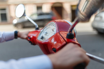 A man driving a red classic scooter rides on the asphalt in the city. Close - up of man's hands holding on to the red scooter's gas handles