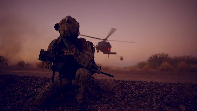 A soldier kneels as helicopters land on a mission behind him.