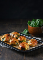 Homemade recipe of croissants stuffed with spinach and ricotta on baking paper in baking dish with bowl of fresh spinach and pine nuts. Dark rustic background. Vertical. Copy space. Healthy concept