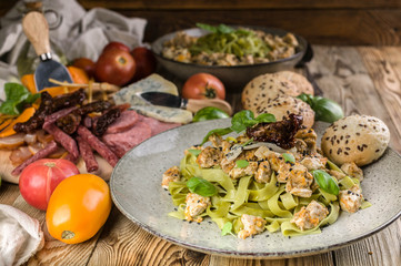 Pesto pasta with meat on a plate, a variety of cheeses and smoked meat and sausage, vegetables and spicy herbs on a wooden table. Close-up