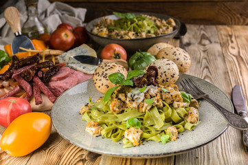 Pesto pasta with meat on a plate, a variety of cheeses and smoked meat and sausage, vegetables and spicy herbs on a wooden table. Close-up