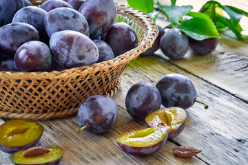Obraz premium Fresh plums with leaves in basket on wooden background