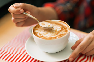 Woman hand with tea spoon over a big cup of a cappuccino coffee with brown cinnamon, cocoa or...
