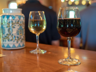 A glass of red and white wine sitting on bar counter top with bartender in back 
