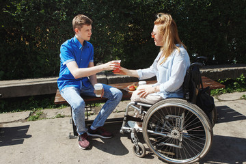 Woman in a wheelchair with her boyfriend drinking coffee and eating muffins in the city's summer park.