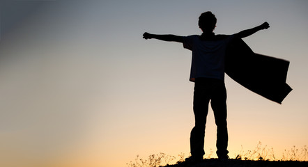 Boy playing superheroes on the sky background, silhouette of teen superhero in a raincoat on the hill - Powered by Adobe