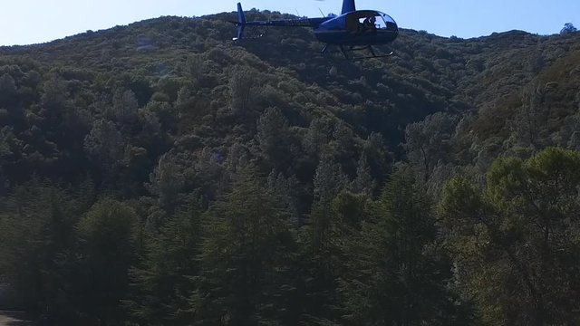 A Robinson R44 helicopter takes off from a mountainous location against a forested backdrop.