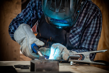 Close up of a young  man welder in  uniform, welding mask and welders leathers, weld  metal  with a arc welding machine  in workshop, blue and orange  sparks fly to the sides