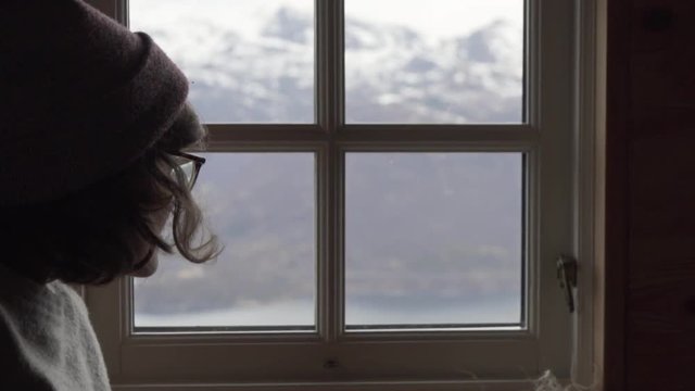 Woman with glasses, grey sweater and red cap inside a wooden cabin leaning towards the window, observing the view of the fjord and snowy mountains and pointing with index finger_slomo