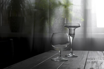 two empty glasses placed on a wooden table on the background of a window with a white curtain. Natural light from a window in a dark room with a flower in the pot. Copy space