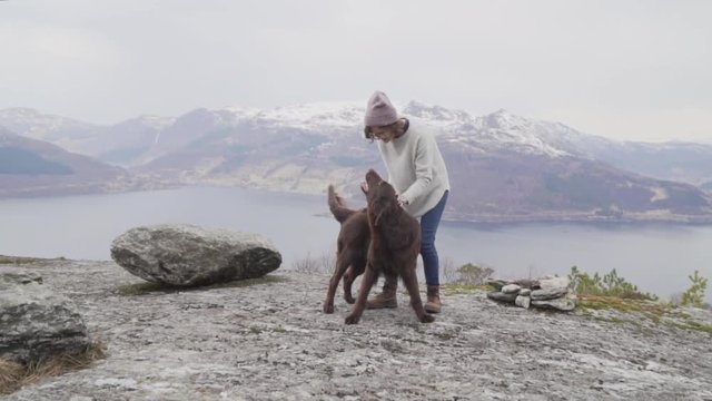 Woman with glasses, red cap, grey sweater, blue jeans and brown boots playing and petting a chocolate flat coated retriever on the top of a mountain with a view to the fjord and snowy mountains