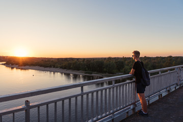teenager guy in life style clothes standing near railings on bridge looking at sunset