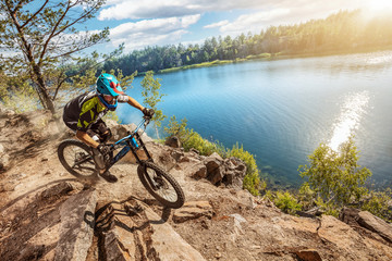 Naklejka na ściany i meble Mountain biker on forest trail near the lake. Male cyclist rides the rock