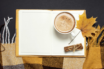 Opened notebook, cup of pumpkin cocoa or coffee, cinnamon, yellow autumn maple leaf, orange brown checkered plaid on black background top view copy space. Autumn flat lay background