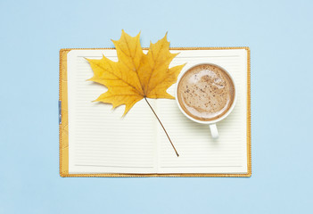Opened notebook, cocoa latte or coffee in cup, yellow autumn maple leaf on blue background top view flat lay. Autumn or winter hot drink, concept of study, working table. Space for text