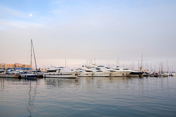 Fototapeta premium Beautiful port of Alicante, Spain at Mediterranean sea. Luxury yachts, ships, ferries and fishing boats sailing and standing in rows in harbor. Rich people traveling around the world. Sunset evening