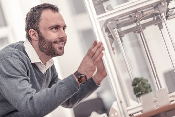 Wonderful thing. Smiling man touching 3D printer standing at the office