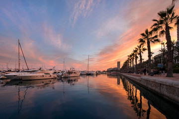 Obraz na płótnie Canvas Beautiful port of Alicante, Spain at Mediterranean sea. Luxury yachts, ships, ferries and fishing boats sailing and standing in rows in harbor. Rich people traveling around the world. Sunset evening