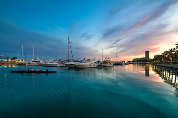 Obraz premium Beautiful port of Alicante, Spain at Mediterranean sea. Luxury yachts, ships, ferries and fishing boats sailing and standing in rows in harbor. Rich people traveling around the world. Sunset evening