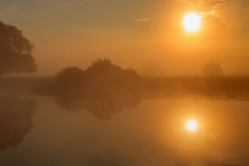 Water surface of river in sunny summer morning with fog and reflection of orange sun. River landscape at sunrise