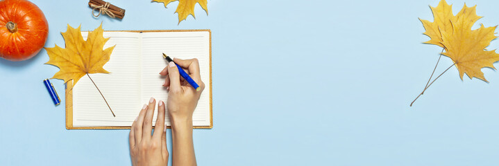 Female hands write in the Open blank notebook with a yellow autumn maple leaf, orange pumpkin, cinnamon on blue background top view flat lay. Autumn concept, study, working table workspace long banner