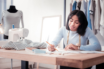 Initial phase. Optimistic qualified dressmaker sitting at the table in her personal studio while...