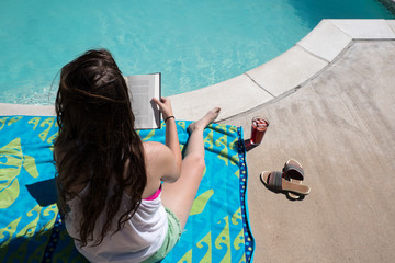 Young brunette woman in shorts and tank top sitting on beach towel reading a book by the swimming pool on a hot summer day. Glass with fruit punch and ice and sandals. Girl reading by the pool.