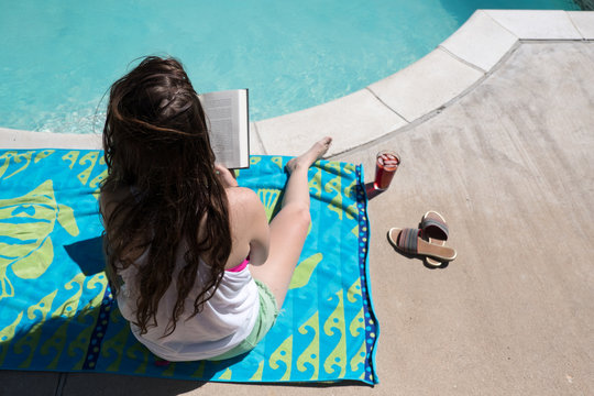 Looking Down On Woman Sitting On Beach Towel Reading A Novel With Her Sandals Off. Brunette In Tank Top And Shorts Reading A Book By The Swimming Pool In The Sun Outside In The Summer.