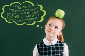 pensive little red haired schoolgirl with apple on head standing near chalkboard with words on different languages in speech bubble