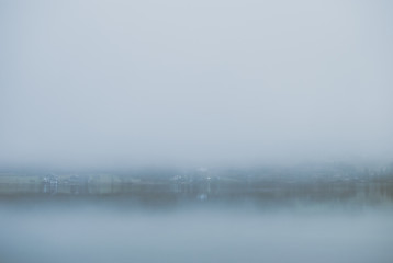 Mysterious mountain lake in early spring morning. Cottages and villas on the other side of lake seen through spooky dense fog covering the water.