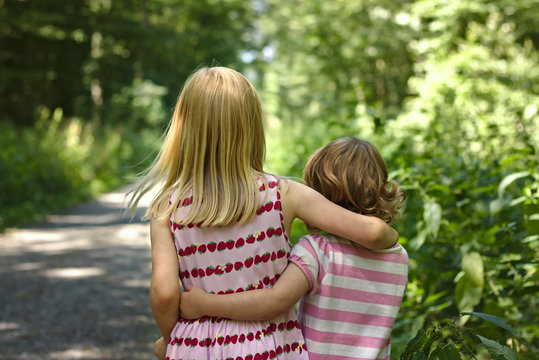 Two little girls wearing summer dresses walk arm in arm along a sunny forest trail