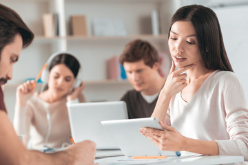 Smart student. Thoughtful Asian woman touching her chin while looking at the tablet screen