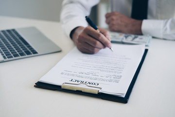 Businessmen holding pen to write business document and contract sheet while sitting at desk with coffee cup, laptop computer and money in home office, Business and Office concept.