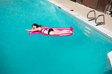 White girl laying on stomach on pink raft. Enjoying lounging in an outdoor swimming pool, blue water and edge of pool. Caucasian girl in bikini on raft in pool arm extended.