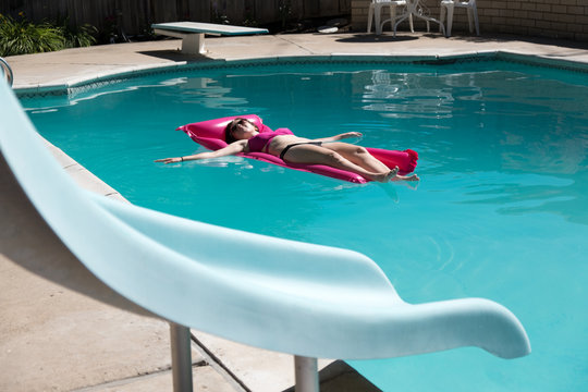 Cute young woman in a bikini on a raft in a swimming pool, slide in the foreground. Woman relaxing on a raft in a backyard swimming pool with slide and diving board on a sunny summer day.