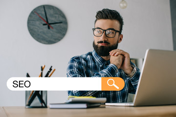 thoughtful young developer looking away while sitting at workplace with laptop and SEO search