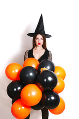 Portrait of beautiful young woman in black witch halloween costume with orange and black balloons over white background