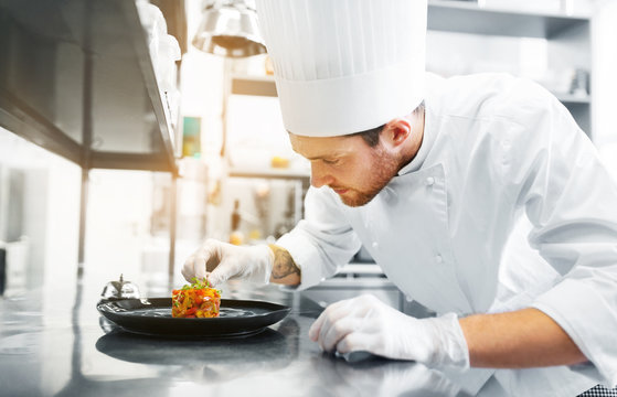 food cooking, profession and people concept - happy male chef cook serving and garnishing stewed vegetables on plate at restaurant kitchen
