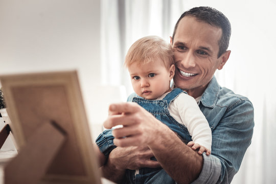 Who is here. Cheerful nice man looking at the photo while showing it to his baby