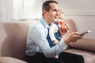 Favourite entertainment. Nice positive man watching TV while staying at home with his baby