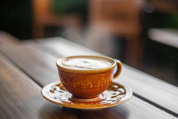 The  brown ceramic cup of hot capucchino coffee on the wooden table in the restaurant or coffee shop with blur people at the background in thw warm yellow light and flare. 