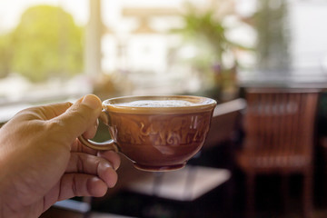 The  white ceramic cup of hot capucchino coffee on the wooden table in the restaurant or coffee shop with blur people at the background in thw warm yellow light and flare. 