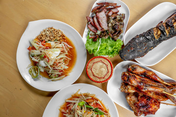 The set of Thai food such as papaya salad , grilled chicken , grilles meat , cat fish and sticky rice in the bamboo container on the wooden table. 