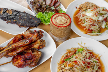 The set of Thai food such as papaya salad , grilled chicken , grilles meat , cat fish and sticky rice in the bamboo container on the wooden table. 