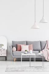 Lamps above table on carpet in white living room interior with pink pillows on grey sofa. Real photo