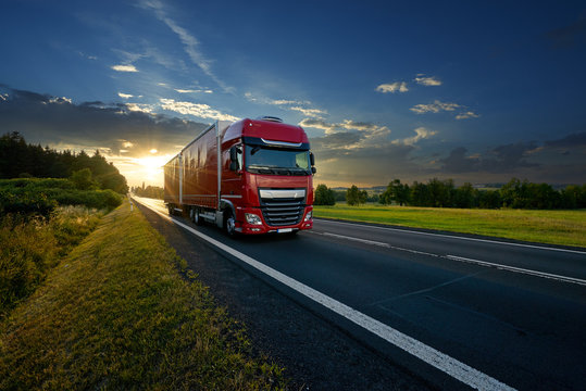 Red truck arriving on the asphalt road in rural landscape in the rays of the sunset