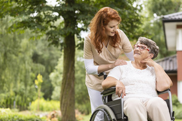 A female volunteer helping an elderly woman in a wheelchair in the garden of an retirement home. - obrazy, fototapety, plakaty