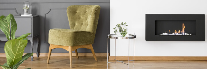 Panorama of a vintage style, comfortable armchair by a gray wall and a silver side table by a black, minimalist fireplace in a classy living room interior