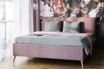 Grey cushions on pink bed in bright hotel bedroom interior with flowers wallpaper. Real photo