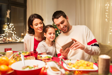 holidays, family and technology concept - happy mother, father and little daughter with smartphone having christmas dinner at home